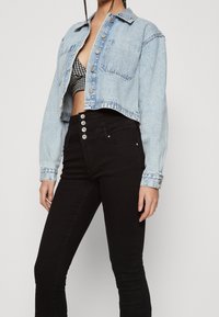 Chaqueta de mezclilla corta de color azul claro con cuello, botones frontales y dos bolsillos en el pecho, combinada con unos pantalones negros ajustados de tiro alto que cuentan con múltiples cierres de botones.