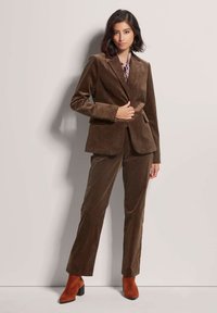 Brauner Cordanzug mit einer taillierten Blazerjacke und geradem Beinform. Kombiniert mit rostfarbenen Stiefeletten. Glatte Textur, dezentes Rippmuster.