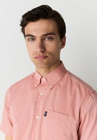 Kortærmet, knappet skjorte i blødt koralfarvet stof. Har en lomme med en lille logo og klassisk knappet skjortekrave.