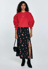 En kvinde med krøllet hår har en rød oversized sweatshirt på og en sort blomsterkjole med en slids til låret, mens hun holder en sort taske.