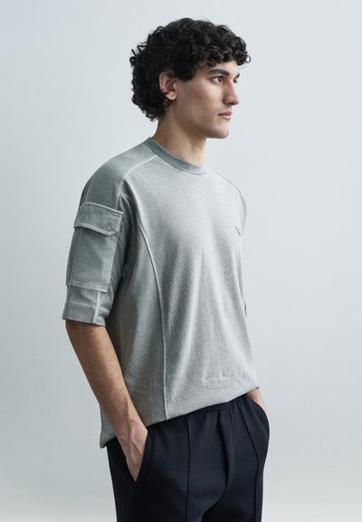 Lysegrå T-shirt med korte ærmer og rund hals, med en lomme på ærmet og tekstureret stof. Kombineret med mørke bukser.