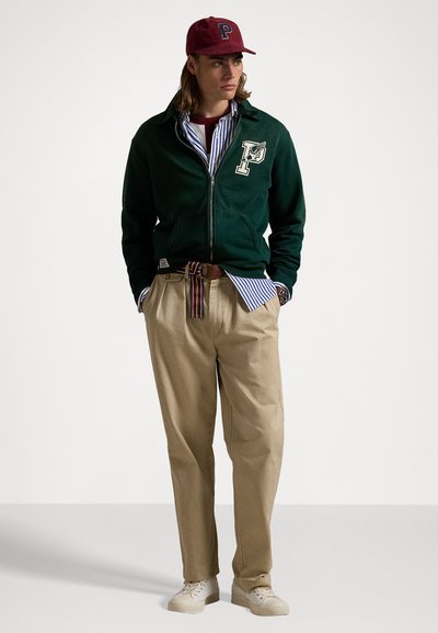 Felpa verde con zip e logo, camicia a righe, pantaloni beige e cappellino bordeaux; sneakers color sabbia completano l'outfit. Tessuto in misto cotone testurizzato.