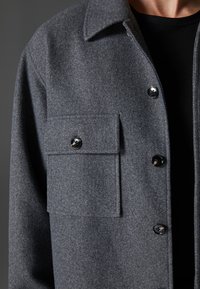 Grå uldfrakke med krave, stort brystlomme og skinnende sorte knapper. Blød tekstur og skræddersyet design.