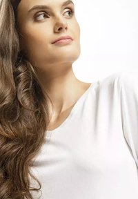 Biała, krótki rękaw bluzka o miękkiej fakturze i luźnym dekolcie; długie, faliste brązowe włosy uzupełniają wygląd na jednolitym tle.