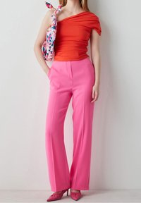 Frau trägt ein rotes Oberteil mit einem Schulterträger, hochtaillierte, weit geschnittene pinke Hose, pinke High Heels und eine Schultertasche mit floralem Muster.