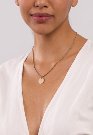 Goldfarbene Kettenhalskette mit einem runden Anhänger mit glatter Oberfläche, getragen über einem weißen V-Ausschnitt-Oberteil vor einem neutralen Hintergrund.