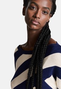 Svetr s námořnickými a krémovými pruhy, který má široký výstřih a texturované pletení. Modelka má dlouhé copánky a neutrální pozadí.