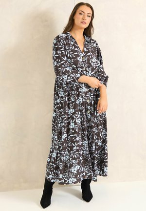 Kobieta poprawiająca rękaw długiej czarnej sukienki z jasnoniebieskim kwiatowym wzorem, w czarnych butach na obcasie, stojąca na neutralnym tle.