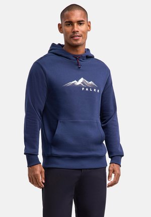 Mężczyzna ubrany w granatową bluzę z kapturem z grafiką góry i napisem „FALKE”, stojący na tle jednolitego jasnoszarego tła.
