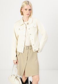 Veste en denim crème avec des fermetures à boutons, des poches poitrine et des manches longues, associée à une jupe ceinturée marron clair et un sac à main.