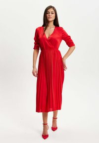 Red pleated dress with a v-neckline and long sleeves, made of satin-like material. Paired with matching red heeled shoes.
