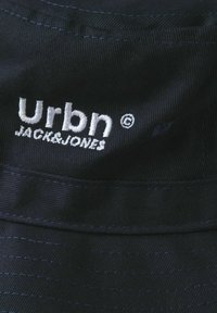 Sort stof bucket hat med hvid broderet logo "Urbn JACK&JONES." Tekstureret materiale med synlig syning på skyggen.