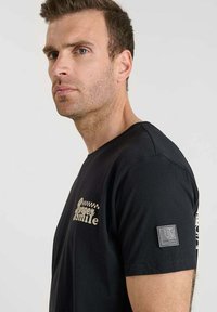 Camiseta negra de algodón, con un gráfico amarillo "Queen Smile" y un parche de logo gris en la manga. Diseño de mangas cortas.