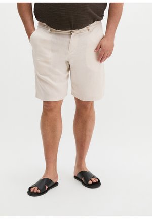 Mann trägt beige Shorts, dunkelbraunes Hemd und schwarze Sandalen mit gekreuzten Riemen, steht mit den Händen nahe den Taschen vor weißem Hintergrund.