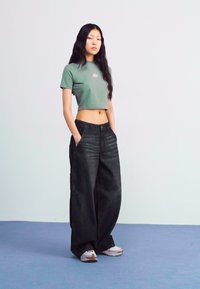 Green cropped t-shirt with a small logo, paired with loose-fitting black denim pants. Model wears white sneakers; minimalistic background.