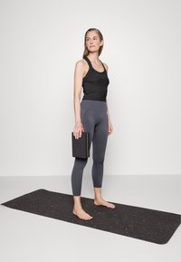 Top preto e leggings cinzentos usados por uma figura em pé segurando um bloco preto, sobre um tapete de yoga escuro com manchas coloridas.