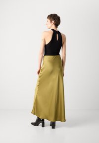 Black sleeveless top with a keyhole back, paired with a long, flowing olive green satin skirt and black block-heeled ankle boots.