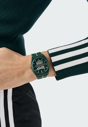 Orologio Adidas verde scuro al polso con il testo "Summer League" sul quadrante, abbinato a manica a righe nere e bianche e pantaloni coordinati.