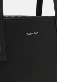 Sac à main noir en cuir lisse, avec un design minimaliste et le logo Calvin Klein argenté sur le devant. Détail zippé en haut.