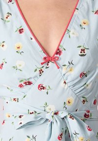 Rochie florală de culoare bleu deschis, cu decolteu adânc în V, finisaje roșii, detaliu cu fundiță mică și modele florale vibrante în roșu, galben și alb.