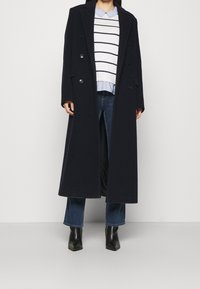 Long manteau en laine bleu marine foncé avec des boutons croisés, superposé sur un pull rayé et un jean, complété par des bottines noires.