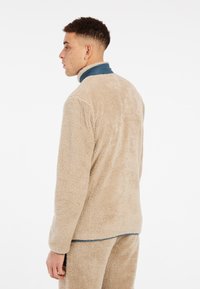 Giacca in pile beige con una texture morbida, collo blu a contrasto e vestibilità rilassata. Presenta una finitura liscia e un orlo leggermente curvo.