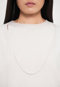 Nærbillede af en person iført en lys beige top med en unik udskæring, der har udsnit ved skuldrene.