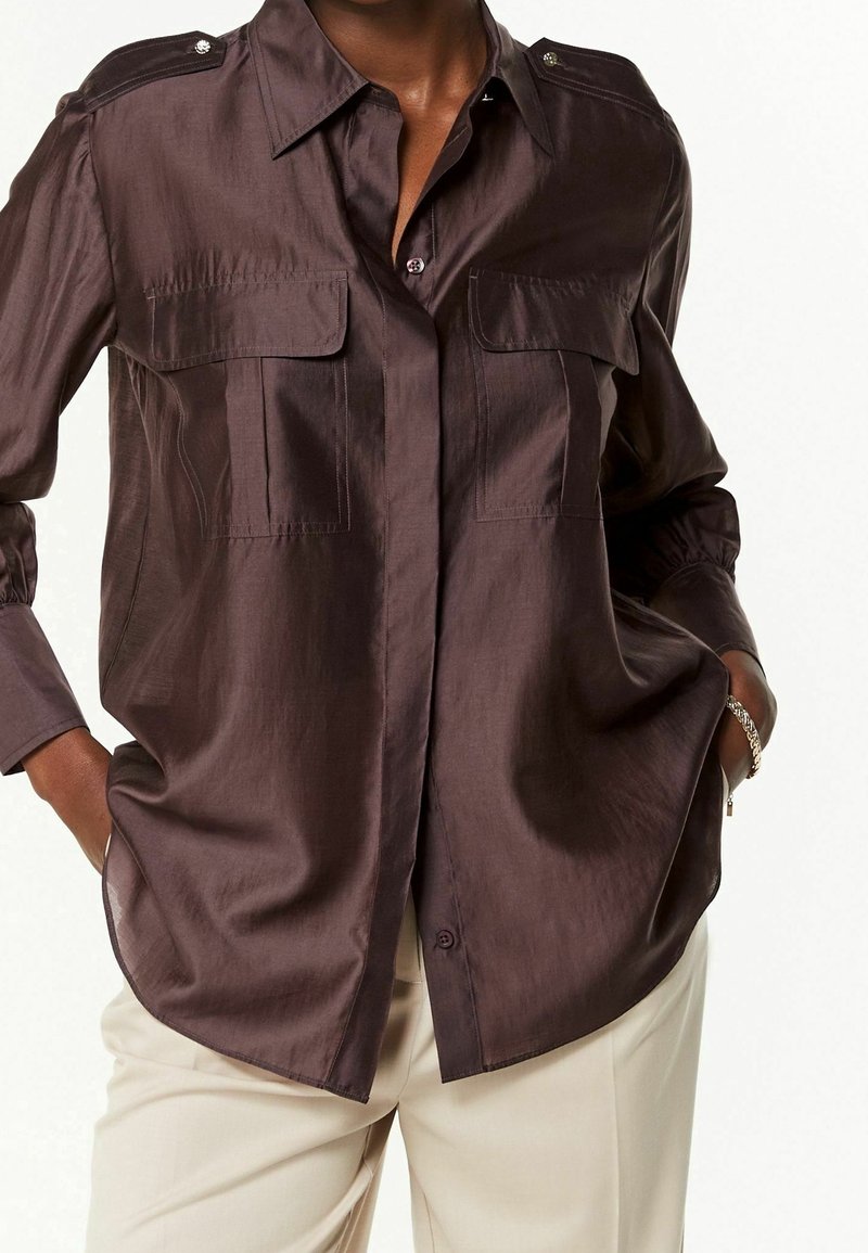 Blouse en satin bronze avec deux poches poitrine, fermeture à boutons et détails aux épaules. Manches longues avec poignets froncés, ourlet légèrement arrondi.