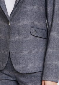 Blå strukturerad blazer med herringbone-mönster, enkel knäppning, och en liten ficka med en märkespräglad metallaccent.