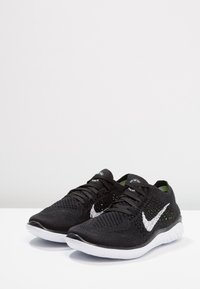 Zwarte sportschoenen met een bovenwerk van mesh, een witte Nike swoosh en groene accenten. Heeft een gestructureerd oppervlak en een witte rubberen zool.