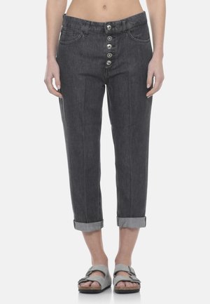 Jeans grigio scuro slim fit con risvolti e bottoni frontali, indossati con sandali chiari con fibbia, su una persona in piedi.