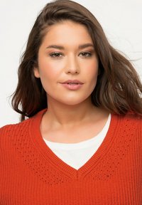 Czerwony sweter dzianinowy z dekoltem w serek i teksturowanym wzorem wokół dekoltu, noszony na białej koszuli. Miękki materiał; luźny krój.