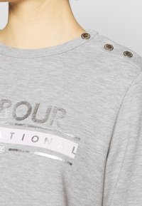 Gray cotton t-shirt featuring metallic silver text and a pink stripe. Notable details include four metallic shoulder snaps and a round neckline.