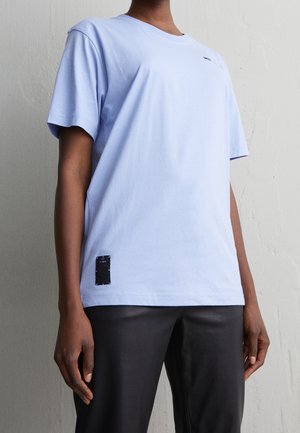 Personne portant un t-shirt à manches courtes bleu clair avec un petit patch noir près de l'ourlet et un pantalon noir, devant un mur uni.