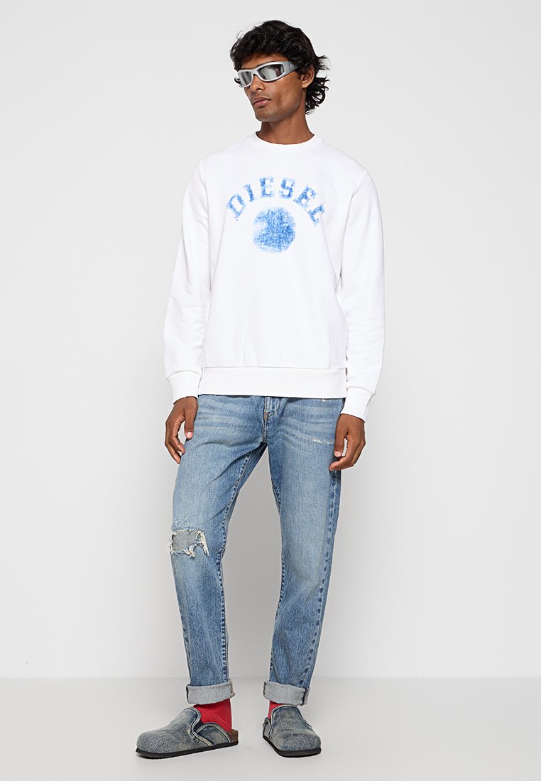 Felpa bianca con logo "DIESEL" blu, abbinata a jeans chiari strappati, scarpe slip-on grigie e calzini grigio-rossastri.