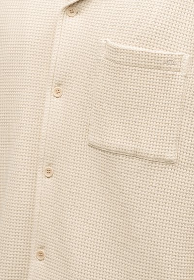Κρεμ υφή πλεκτό ύφασμα με κουμπιά μπροστά και τετράγωνη τσέπη στο στήθος αριστερά.