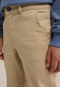 Beige színű, egyenes fazonú pamutnadrág, oldalsó zsebekkel, elöl gombos záródással és sima textúrával, kék pulóverrel párosítva.