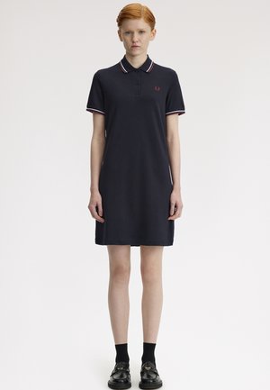 Nuori henkilö, jolla on punaiset hiukset, seisoo ja pukee mustan poolomekon, jossa on raidallinen kaulus ja hihat, mustat sukat ja mustat kävelykengät.