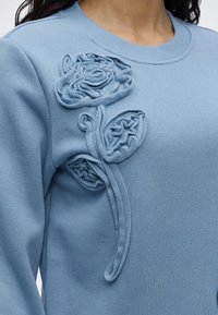 Pull bleu clair avec application florale texturée représentant une rose et des feuilles sur le haut de la poitrine près de l'encolure.
