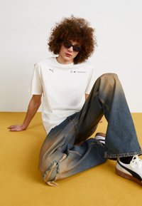 Camiseta blanca de algodón con logo gris, combinada con denim azul desgastado, mostrando un desgarro en la rodilla. Zapatillas blancas y marrones completan el look.