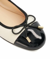 Ballerines en cuir crème avec un bout en vernis noir. Présentent un nœud noir et des accents métalliques dorés. Texture lisse et forme arrondie.