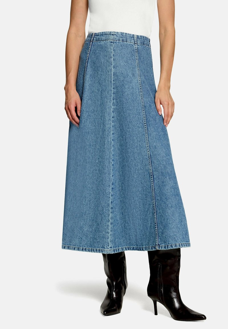 Femme portant une jupe en jean bleu moyen en forme trapèze associée à des bottes en cuir noires à talons hauts et un haut blanc sans manches, debout.