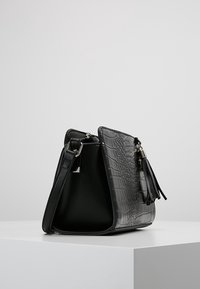 Sort faux læder håndtaske med krokodilletekstur, trapezformet, sølvhardware og en frynseaccent. Indvendigt er den glat og ufodret.