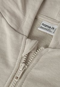 helebeeži kapuutsiga dressipluus, valmistatud pehmest kangast, millel on esiküljel lukuga sulgur, peen tikkimine ning kaeluse siseküljel asuv riietuse silt.