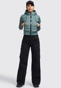 Jachetă puffer verde deschis cu glugă și fermoar în față, asociată cu pantaloni cargo negri cu buzunare mari. Adidașii negri completează ținuta.