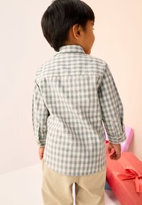 Chemise à manches longues à carreaux verts et blancs avec un poignet boutonné, associée à un pantalon beige clair. En arrière-plan, des boîtes-cadeaux colorées.