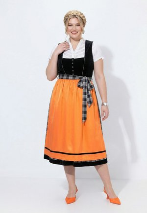 Mujer con el cabello trenzado que lleva blusa blanca, chaleco negro, falda naranja con fajín a cuadros y tacones naranjas a juego, de pie frente a un fondo blanco.