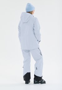 Ljusblå vattentät jacka med huva, kombinerad med matchande byxor och svarta detaljer och dragkedjor. Svarta skidstövlar fulländar outfiten.