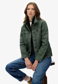 Groene denimjack met twee borstzakken, voorknopen en een hoge kraag. Gecombineerd met blauwe opgerolde jeans en bruine schoenen.