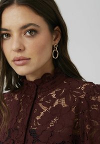 Blouse en dentelle bordeaux avec des motifs floraux et un col montant. Des boucles d'oreilles ovales dorées ajoutent du détail. Teint de peau lisse et couleur de lèvres neutre.